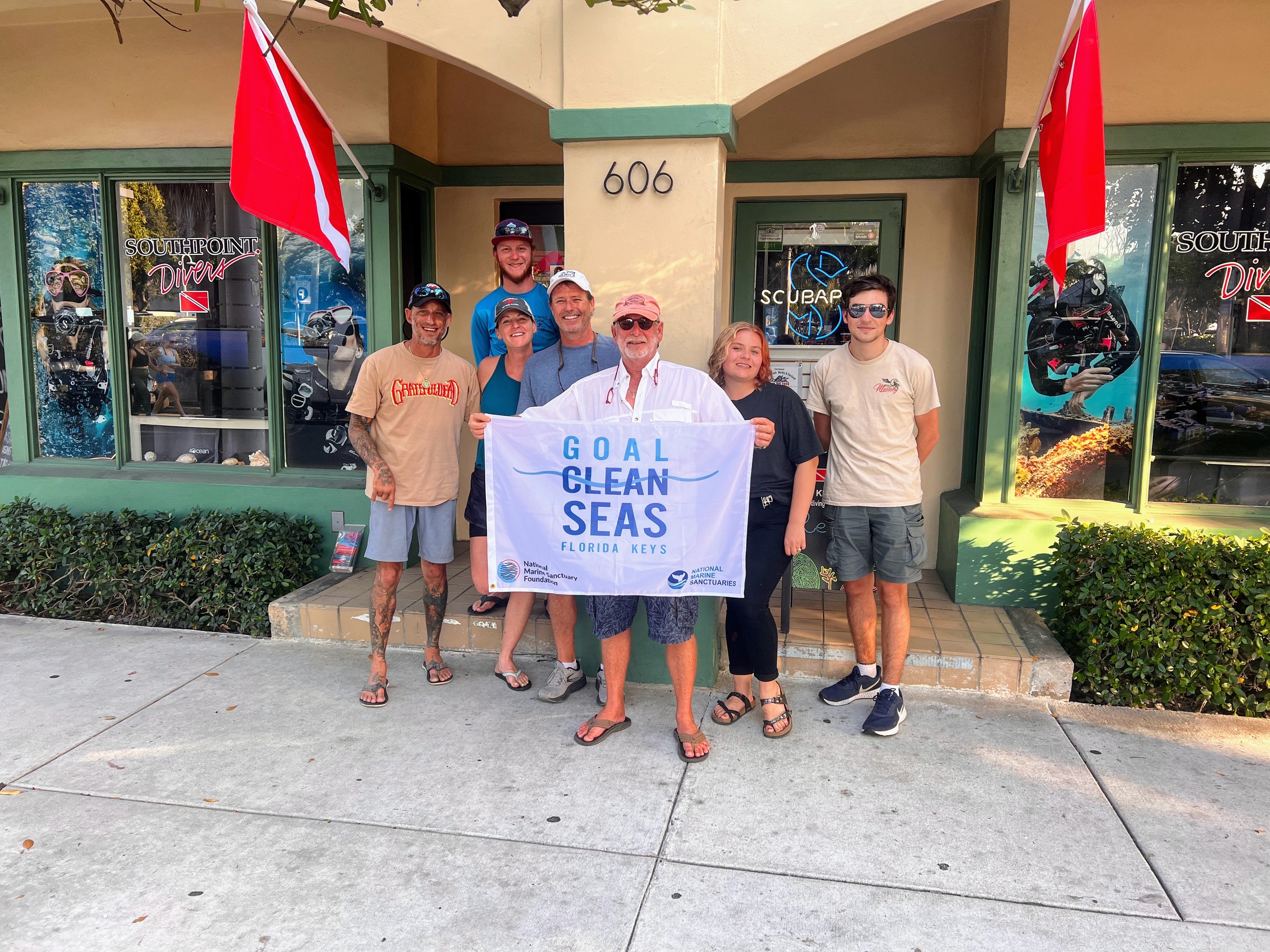 Group holding 'Goal Clean Seas' banner outside dive shop with scuba gear displays.