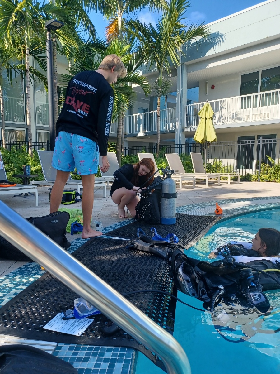 People preparing scuba gear near a hotel pool with palm trees.