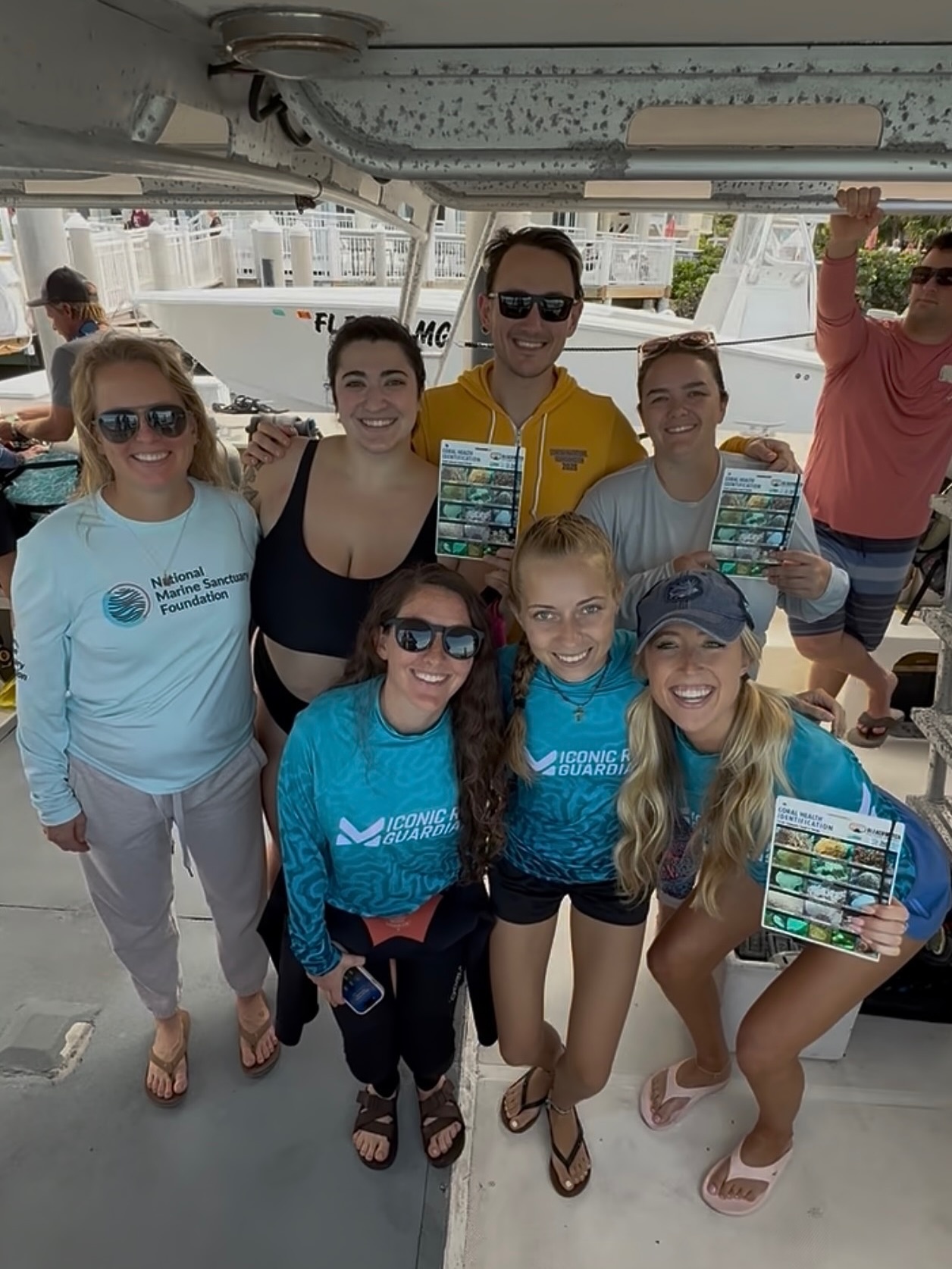 Group of people smiling on a boat, some holding brochures, wearing casual and beachwear.