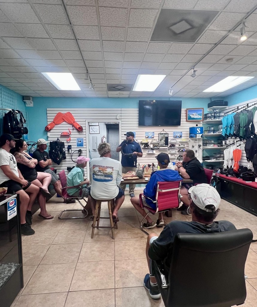 Group of people sitting in a dive shop with a man speaking at the front.