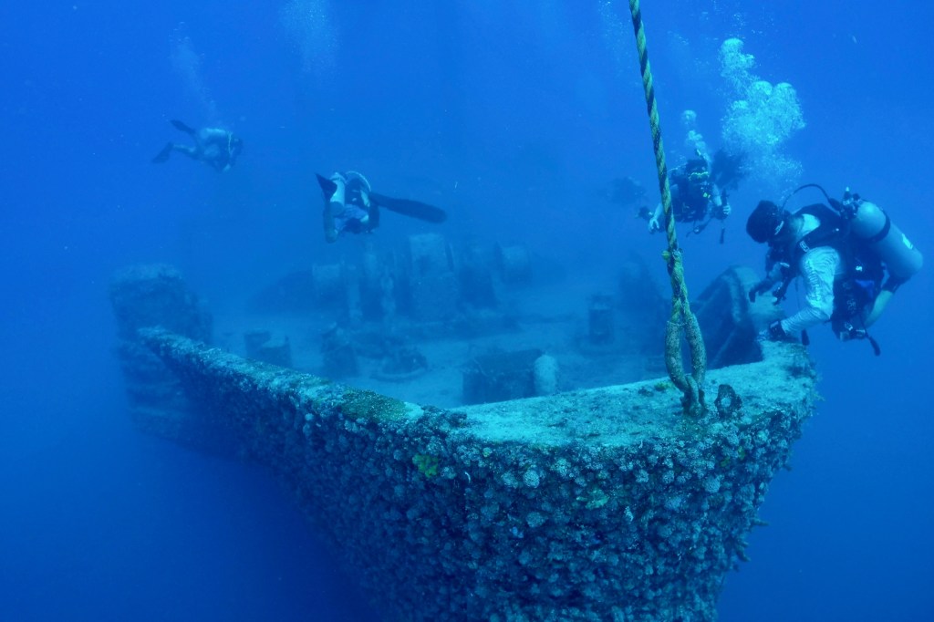 Divers explore a submerged shipwreck with coral growth, surrounded by deep blue water.
