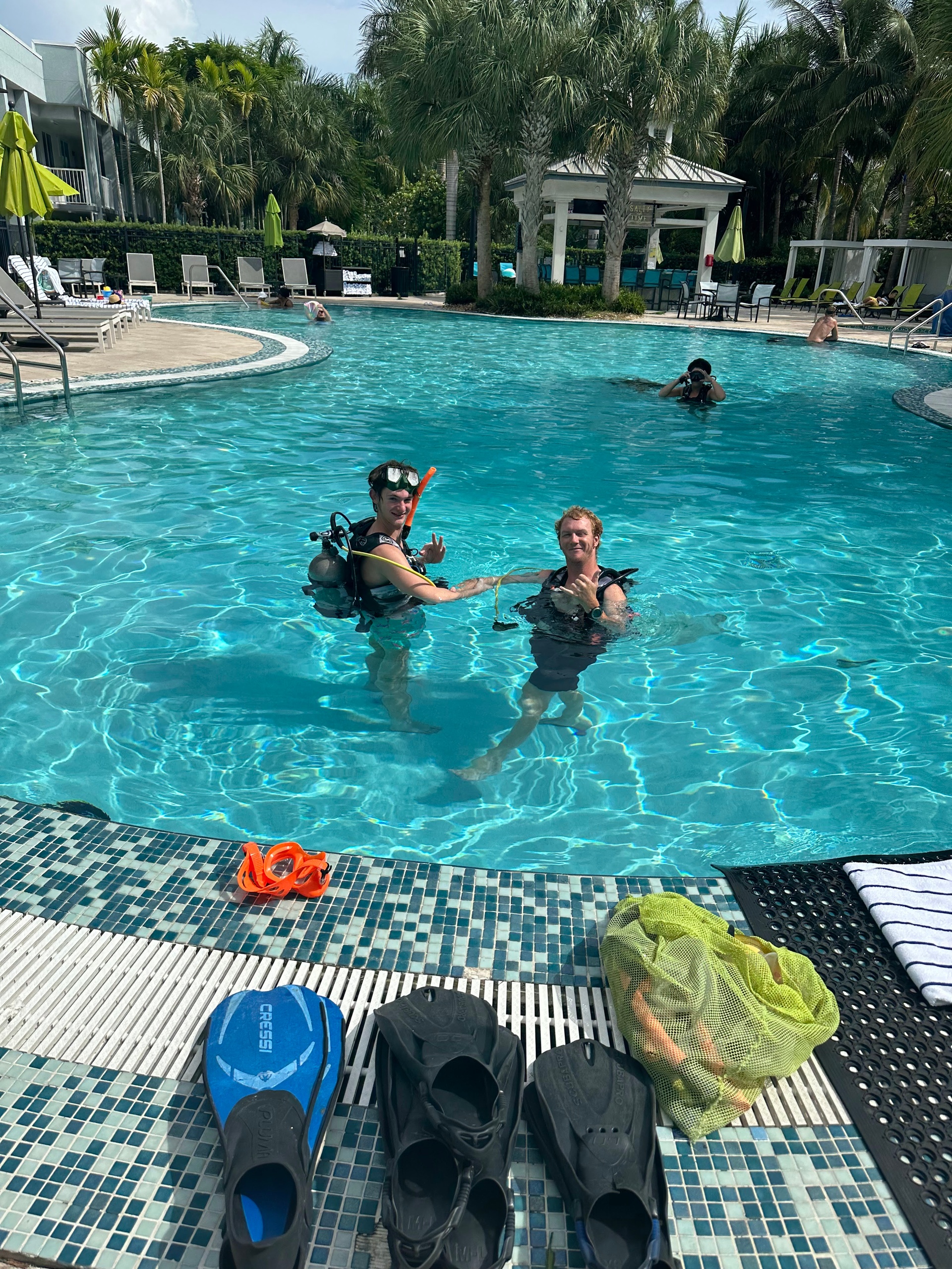 Two divers in a pool near palm trees, swim gear on poolside.