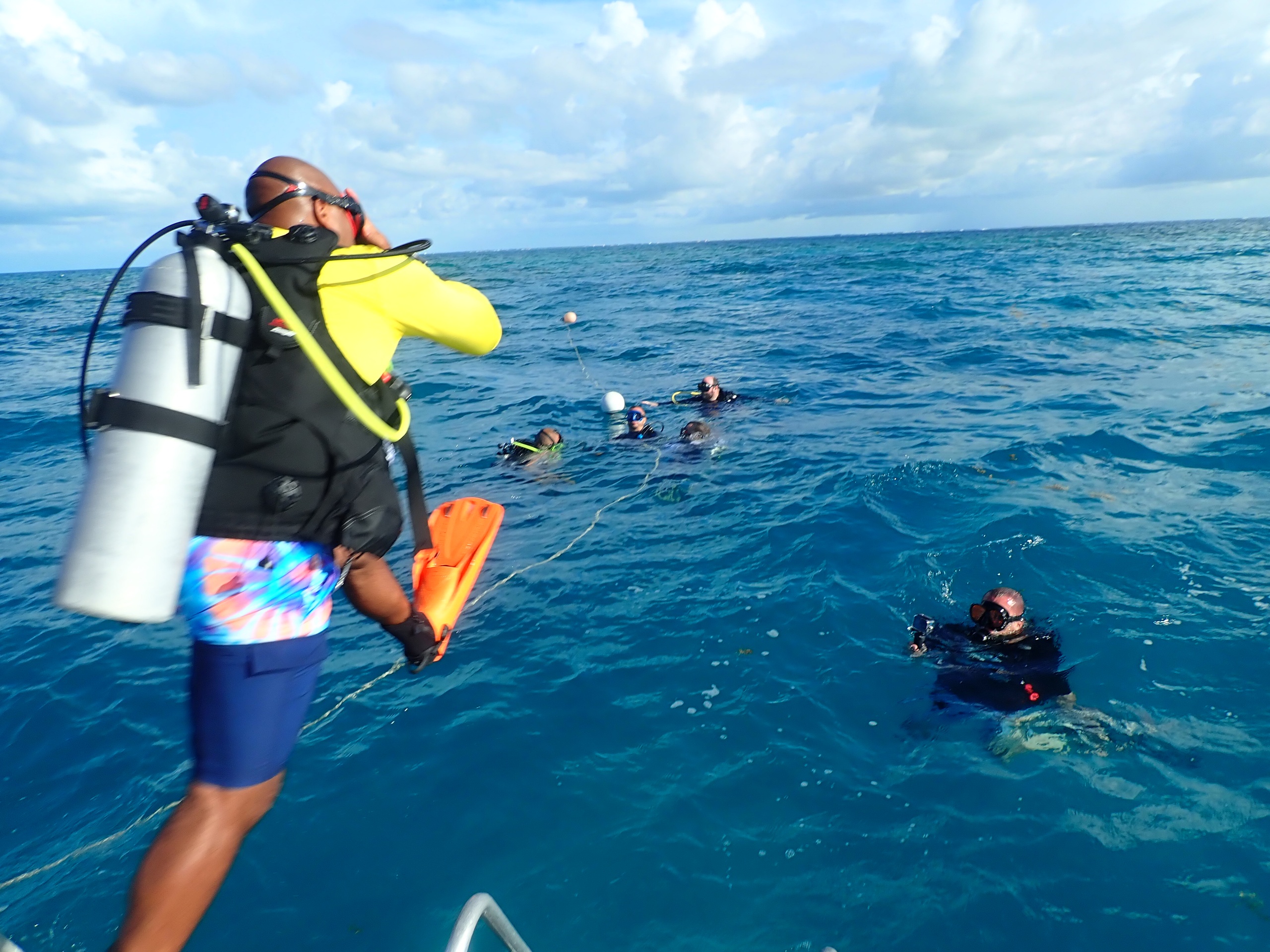 Divers in ocean; one on boat in gear, others in water, cloudy sky.