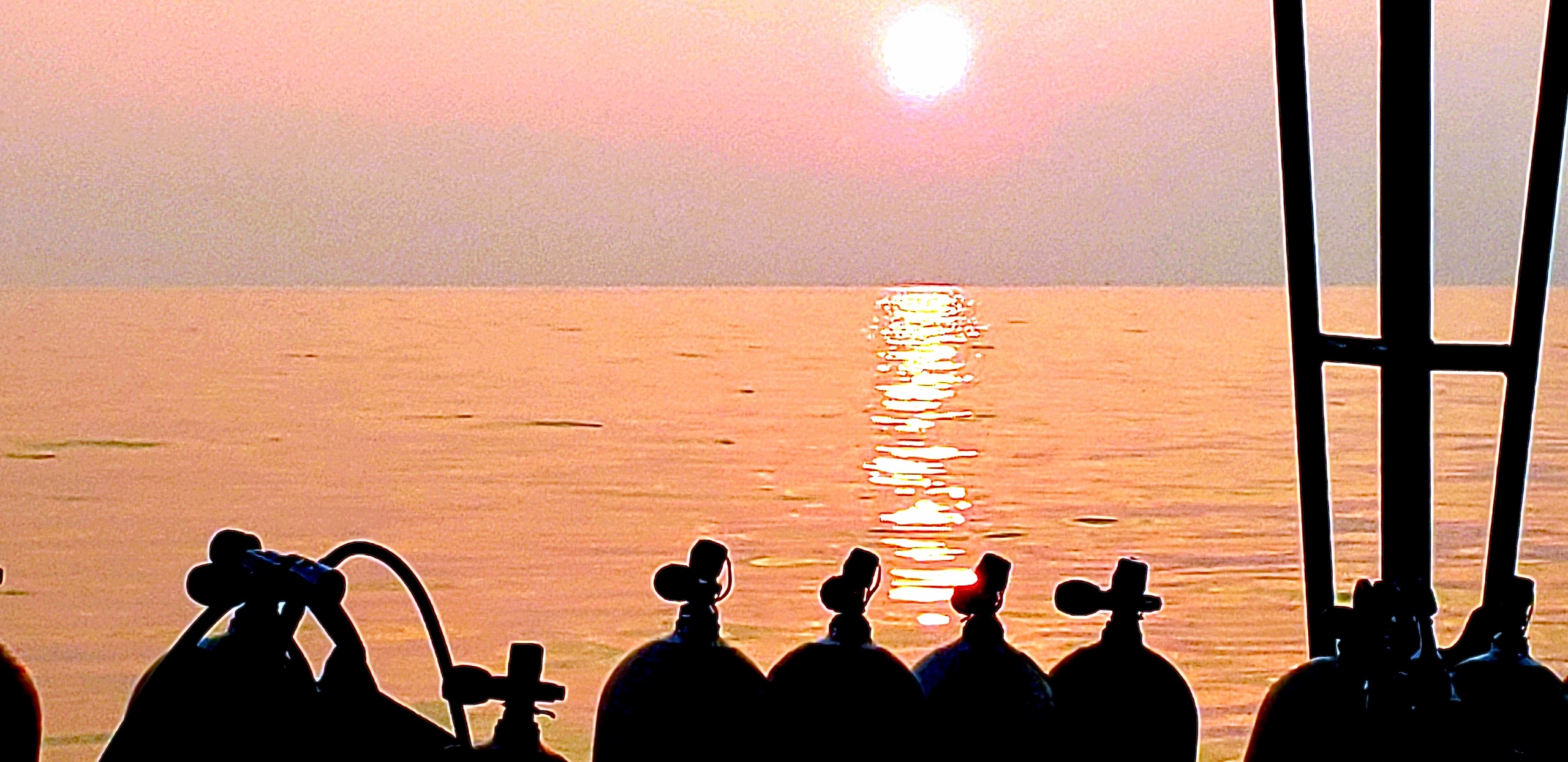 Silhouetted diving tanks on a boat at sunset on a calm sea.