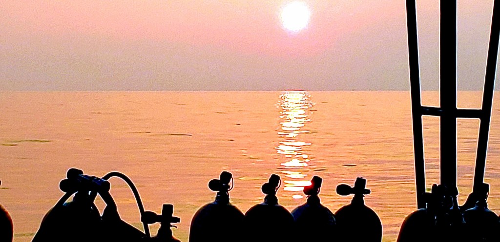 Silhouetted diving tanks on a boat at sunset on a calm sea.