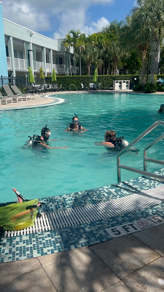 Three people in scuba gear practice in a hotel pool under sunny skies.