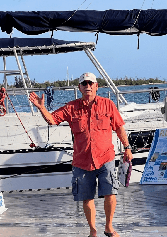 Man in red shirt and cap standing near a sailboat on a sunny day.