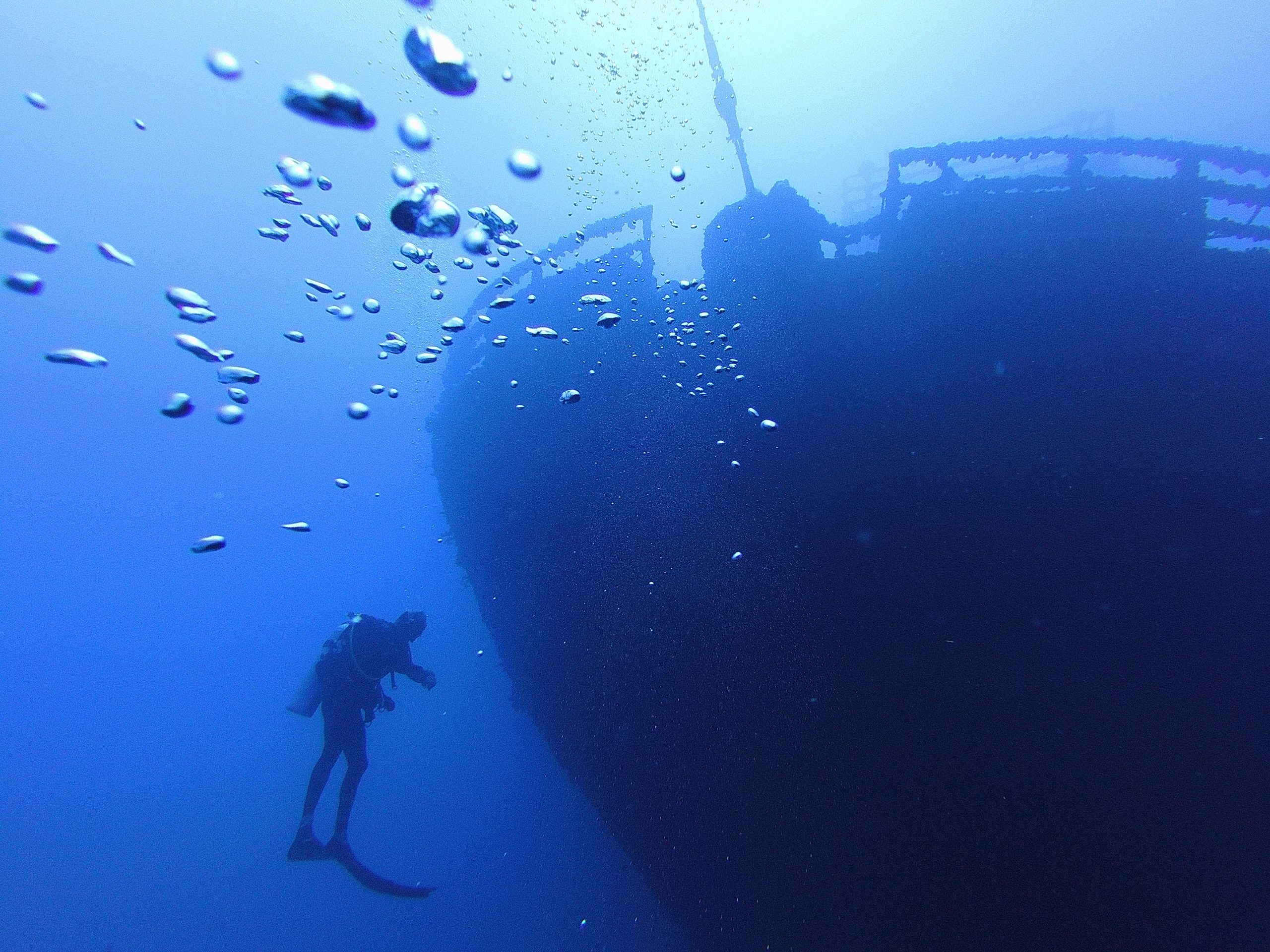Scuba diver near a large sunken ship, with bubbles ascending in blue water.