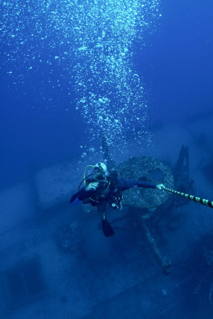 Diver with bubbles ascending near a submerged shipwreck.